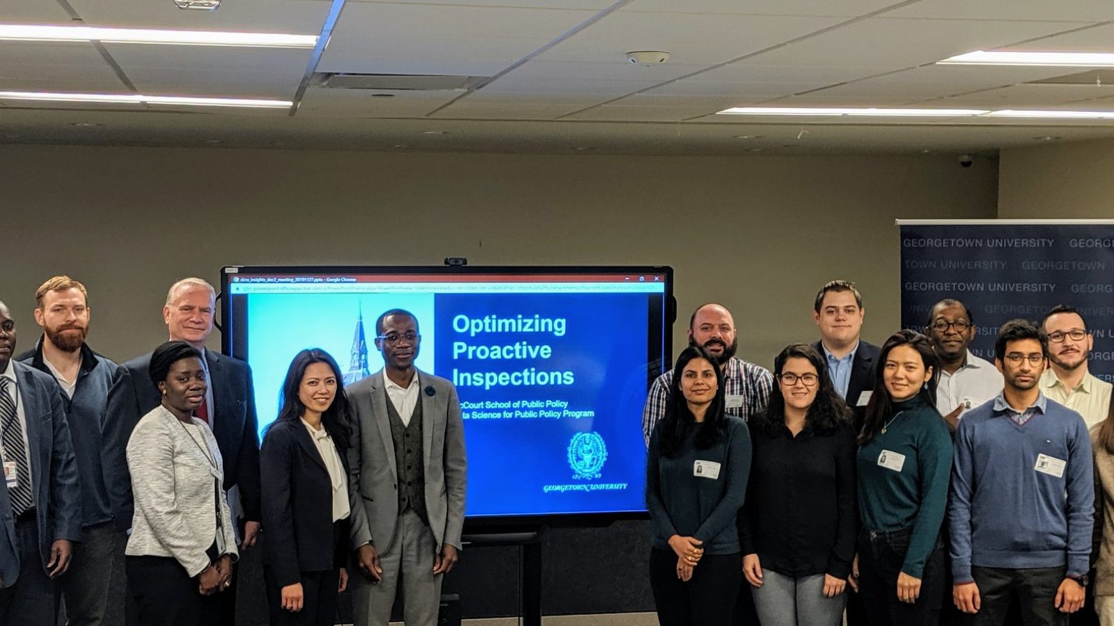 A group of students stand by a TV monitor displaying their presentation &quot;Optimizing Proactive Inspections&quot; to McCourt faculty and the DC Department of Consumer and Regulatory Affairs