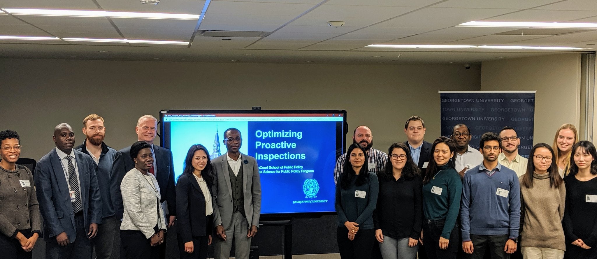 A group of students stand by a TV monitor displaying their presentation "Optimizing Proactive Inspections" to McCourt faculty and the DC Department of Consumer and Regulatory Affairs