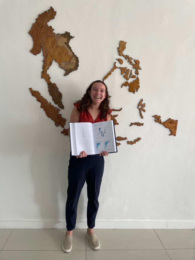 Hannah Reynolds (MIDP'25), posing in front of a map of the Southeast Asia region, proudly presents the heat map and charts she created for a final report on an IDinsight project for the Philippine Department of Health.