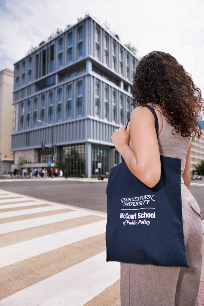 A student in a tan outfit standing in front of the McCourt School of Public Policy holding a navy blue Georgetown McCourt School tote bag