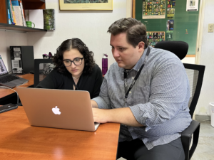 Alec Brockell wearing a plaid button down shirt and black lanyard and Constanza Alarcón wearing a black shirt collaborating on a laptop.