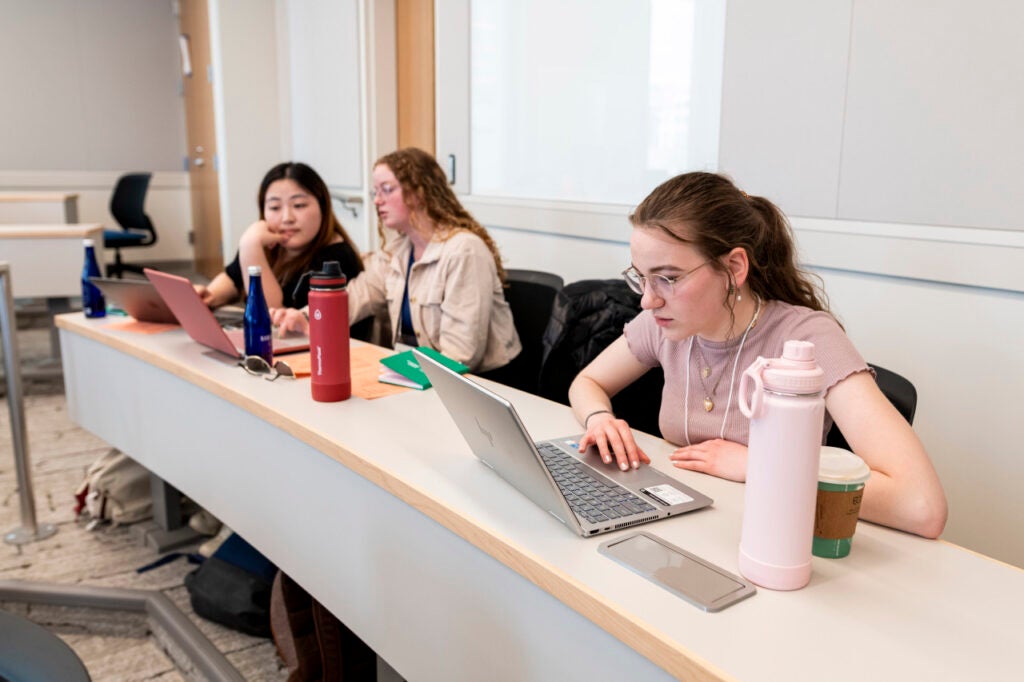 3 students during the McCourt Simulation sitting at a classroom table working on their laptops. 2 students are working together and 1 student is working independently.