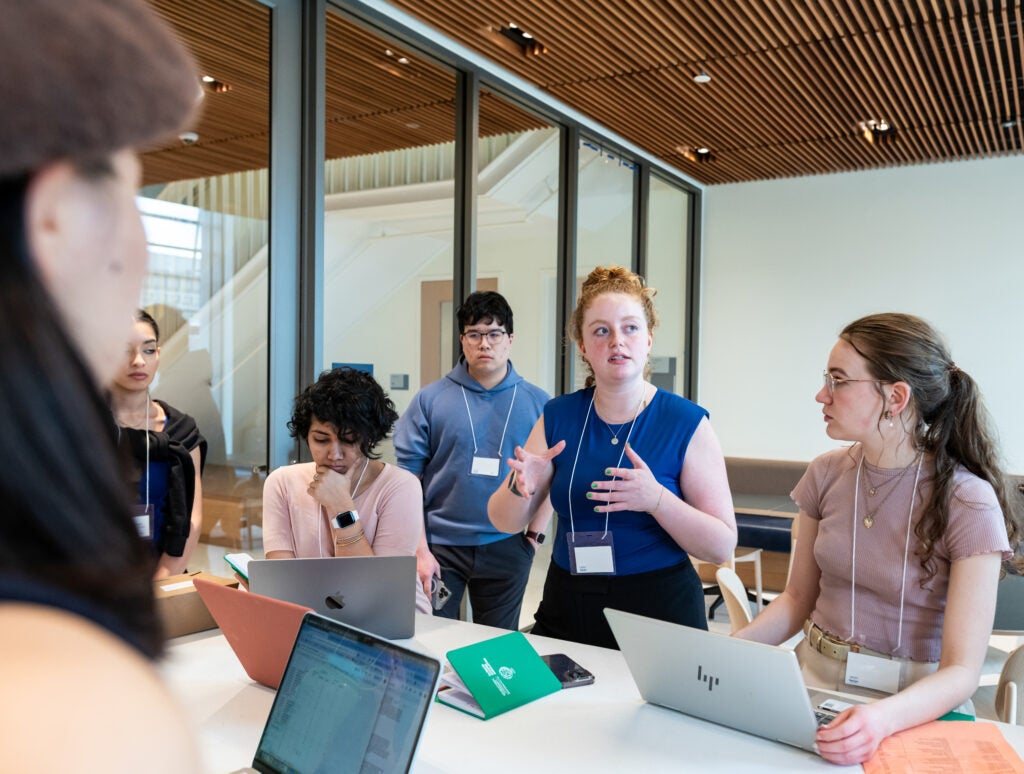 A group of 6 students working on a public policy project together, 1 student is talking with the group while the others work on their laptops or stand and listen