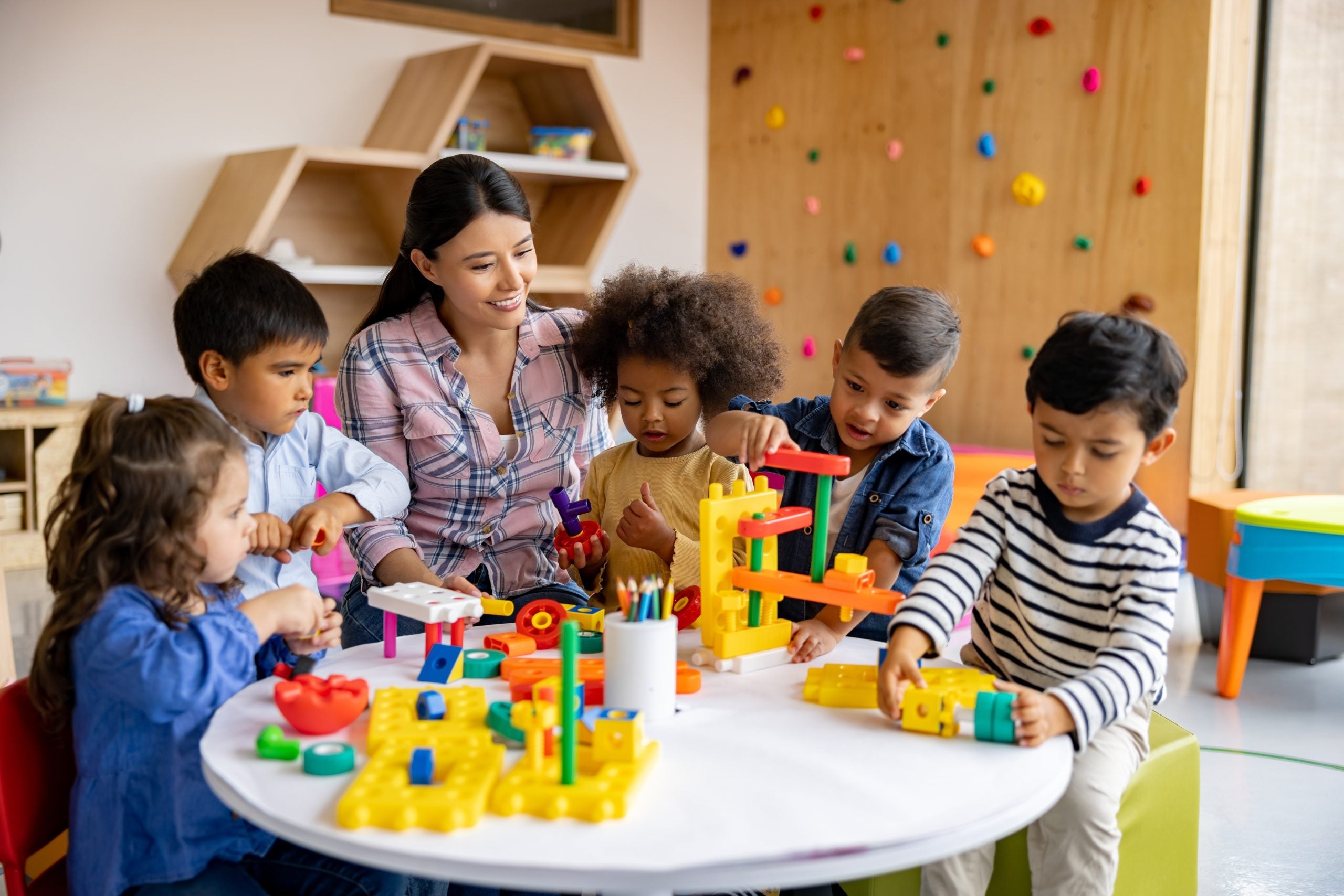 A group of six individuals, five children and one adult, sit at a small table interacting with toys and objects.