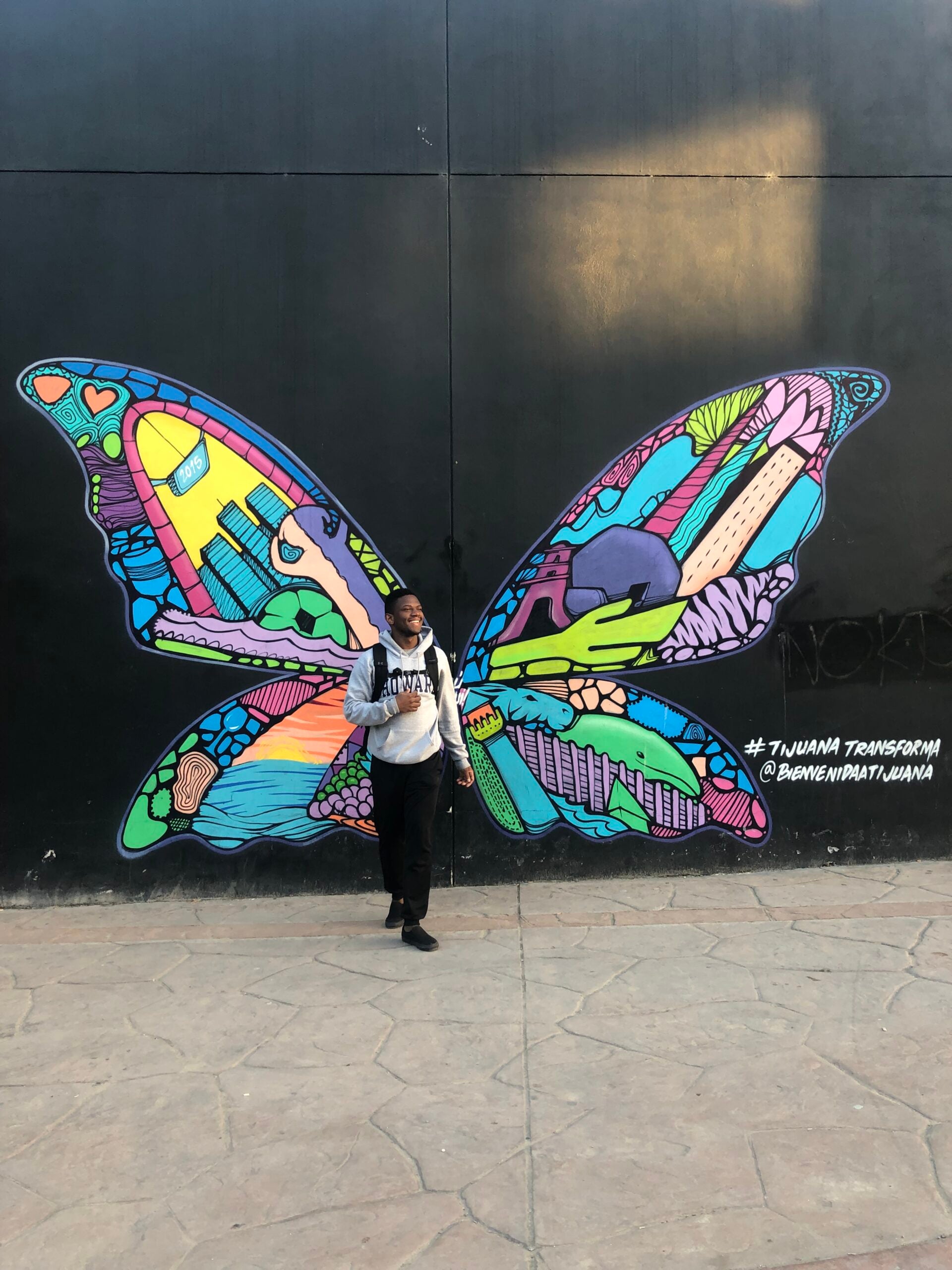 Man standing in front of wall with large and colorful butterfly graffiti as though they were his wings.