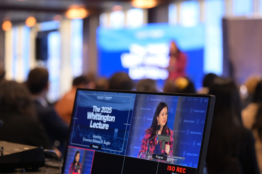 Close-up of production screen showing live footage of Kugler lecturing; background heavily blurred