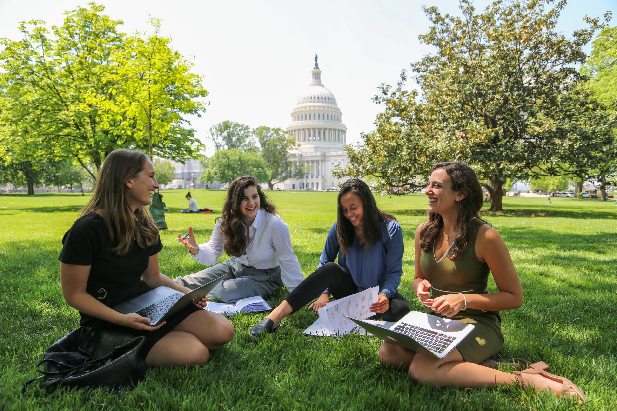 Four people sitting with books and laptops in a grassy field, US Capitol in background