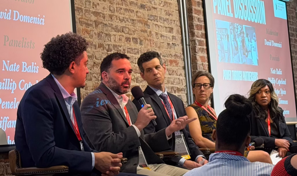 Five people speaking on a panel against a brick wall