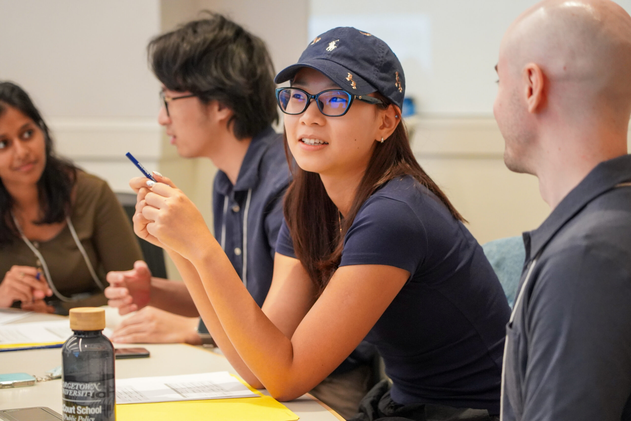 A young woman wearing a navy blue baseball cap and glasses smiles as she speaks, gesturing with a blue pen. 