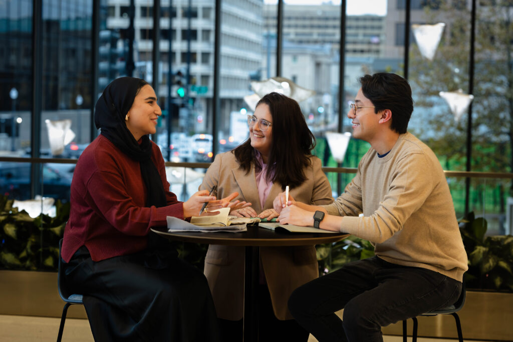 Three students working at a table with books, papers and laptops