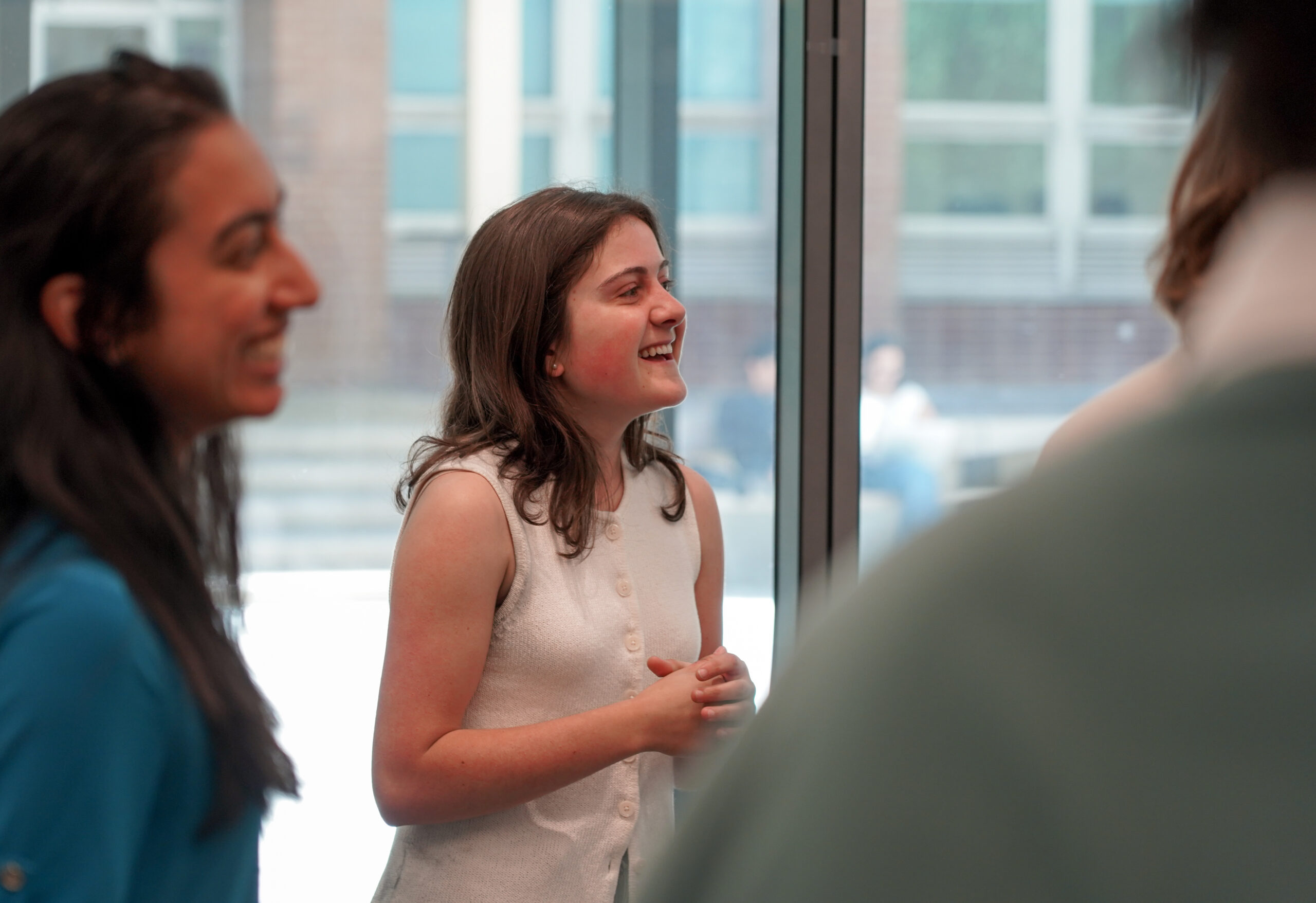 A young woman stands smiling, two other people in foreground heavily blurred