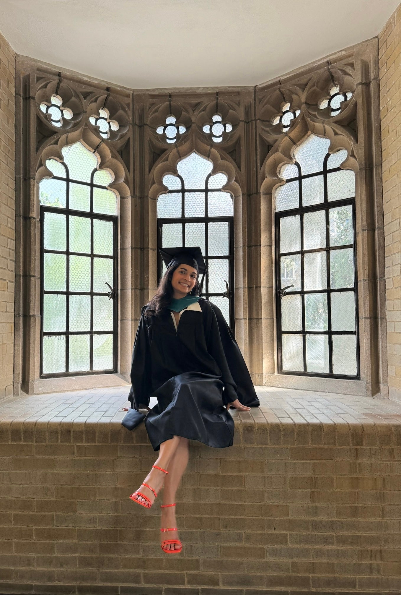 Sneha Choudhary in graduation gown, sitting in front of bay window