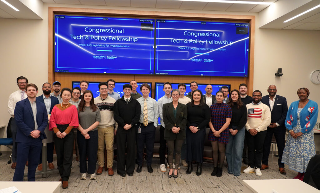 Large group of people standing side-by-side in front of screens reading "Congressional Tech and Public Policy Fellowship"