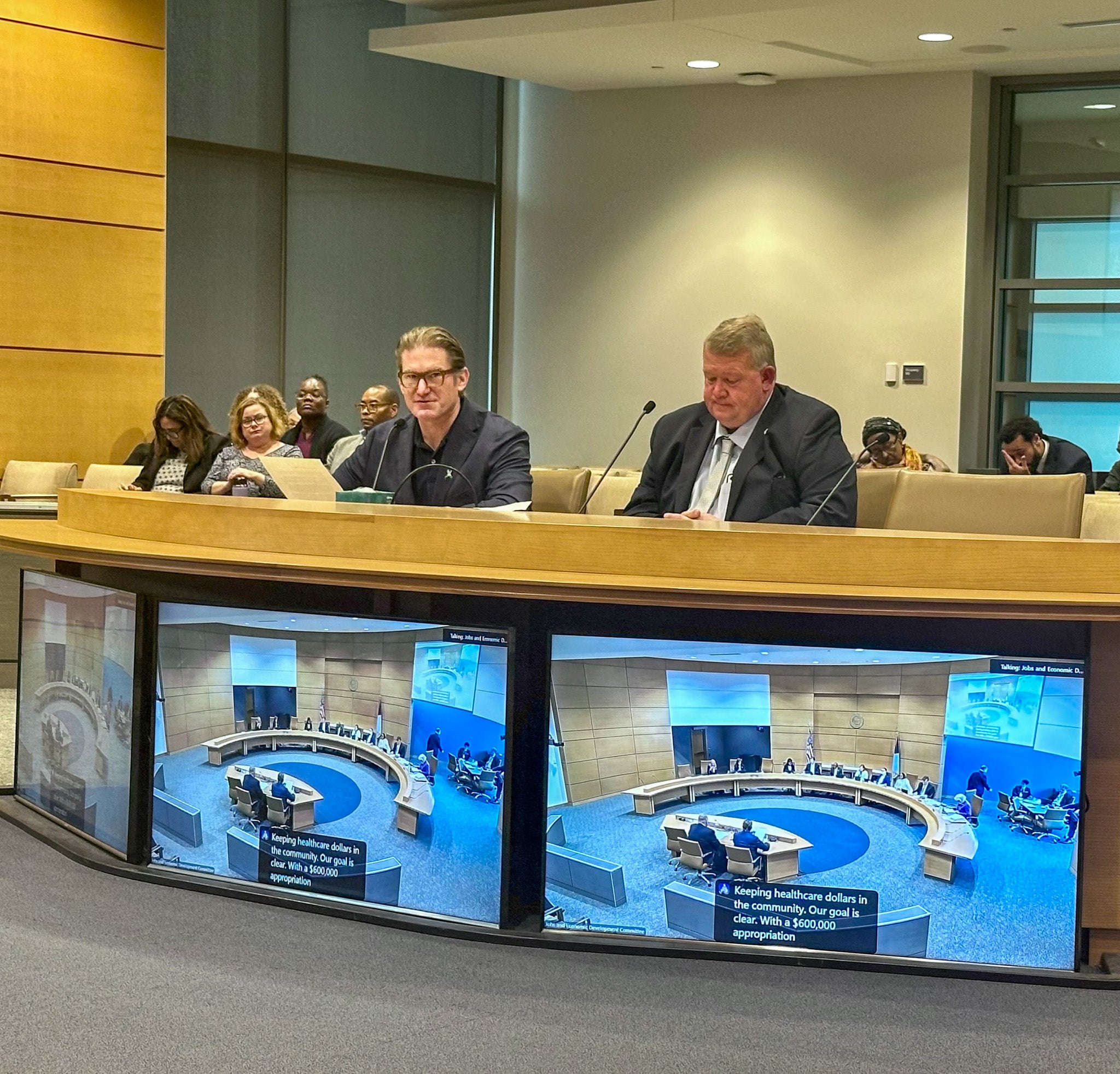 Two individuals sit behind a desk, front of desk is multiple large TV screens showing view of room