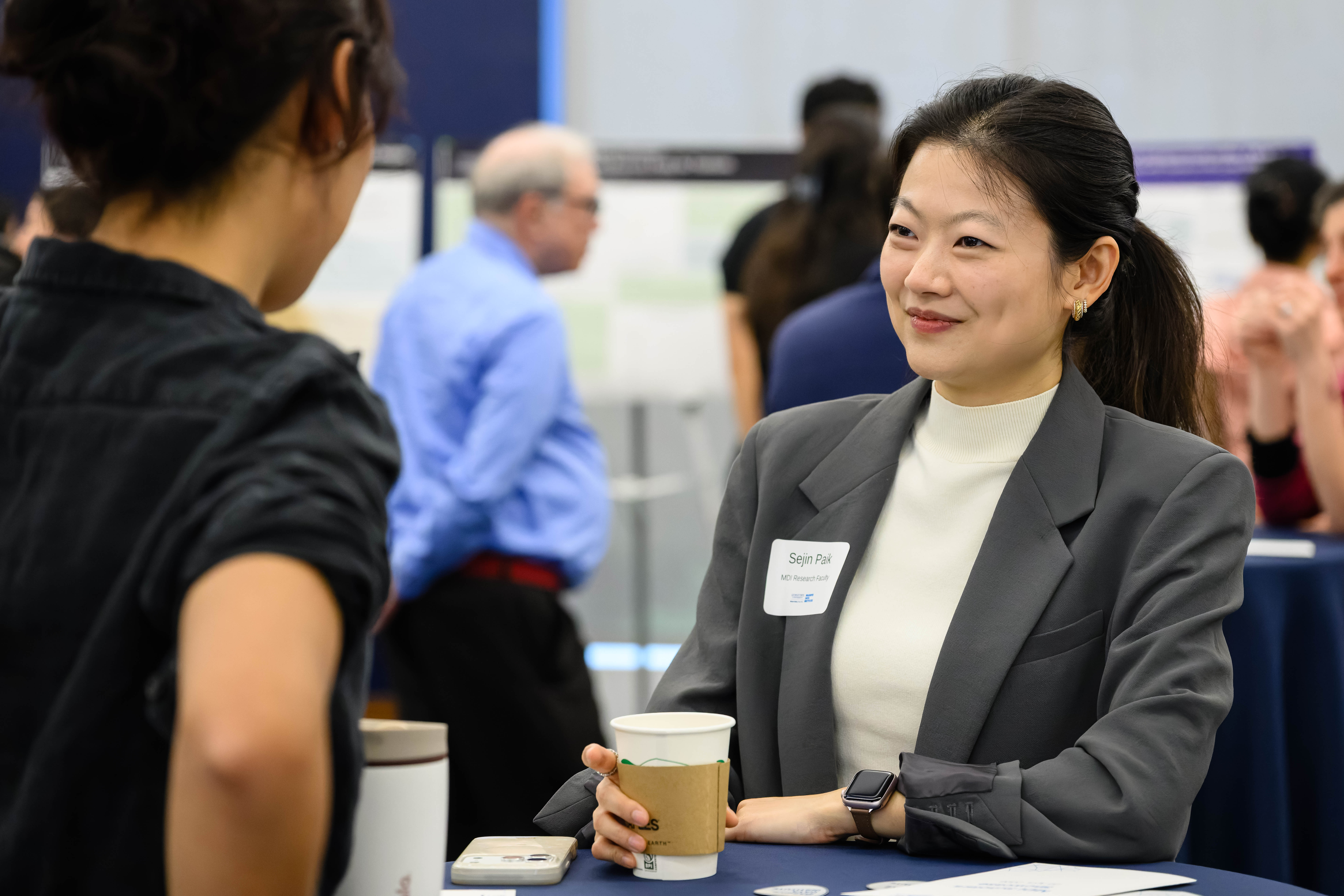 Young asian woman, wearing grey blazer with name badge, speaks to another person
