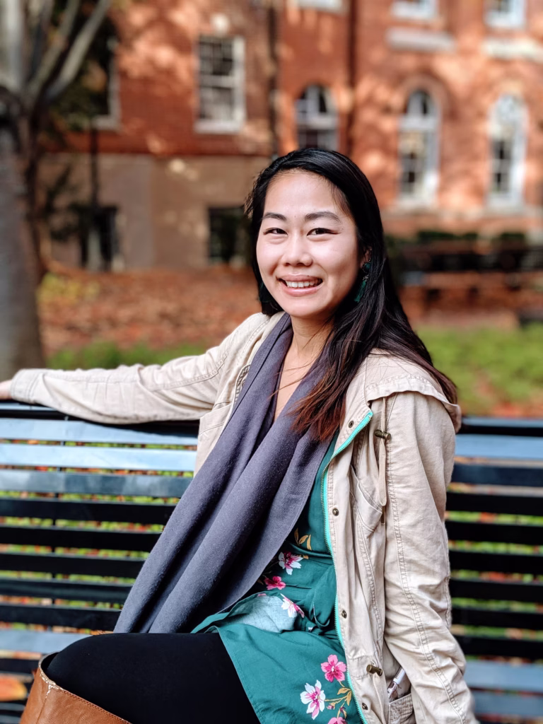 Frances Chen sitting on a park bench wearing a tan jacket, turquoise dress, and black leggings.