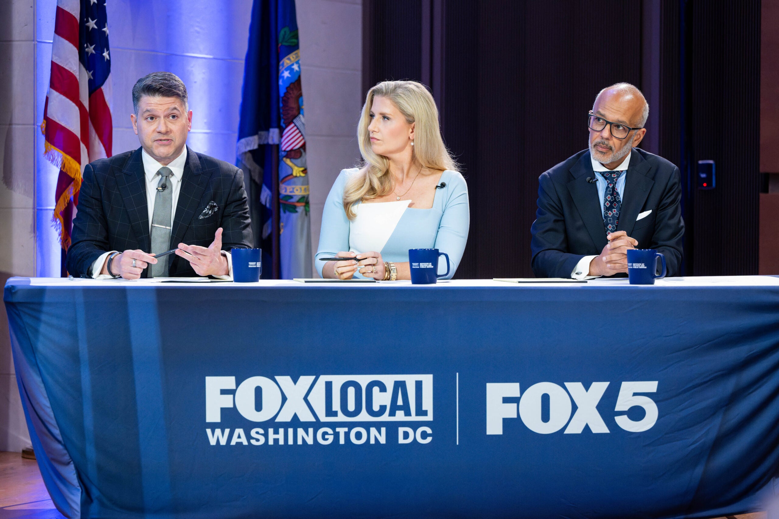 Three people sat side-by-side, dressed professionally, moderating mayoral debate; sat behind desk that reads "Fox 5"