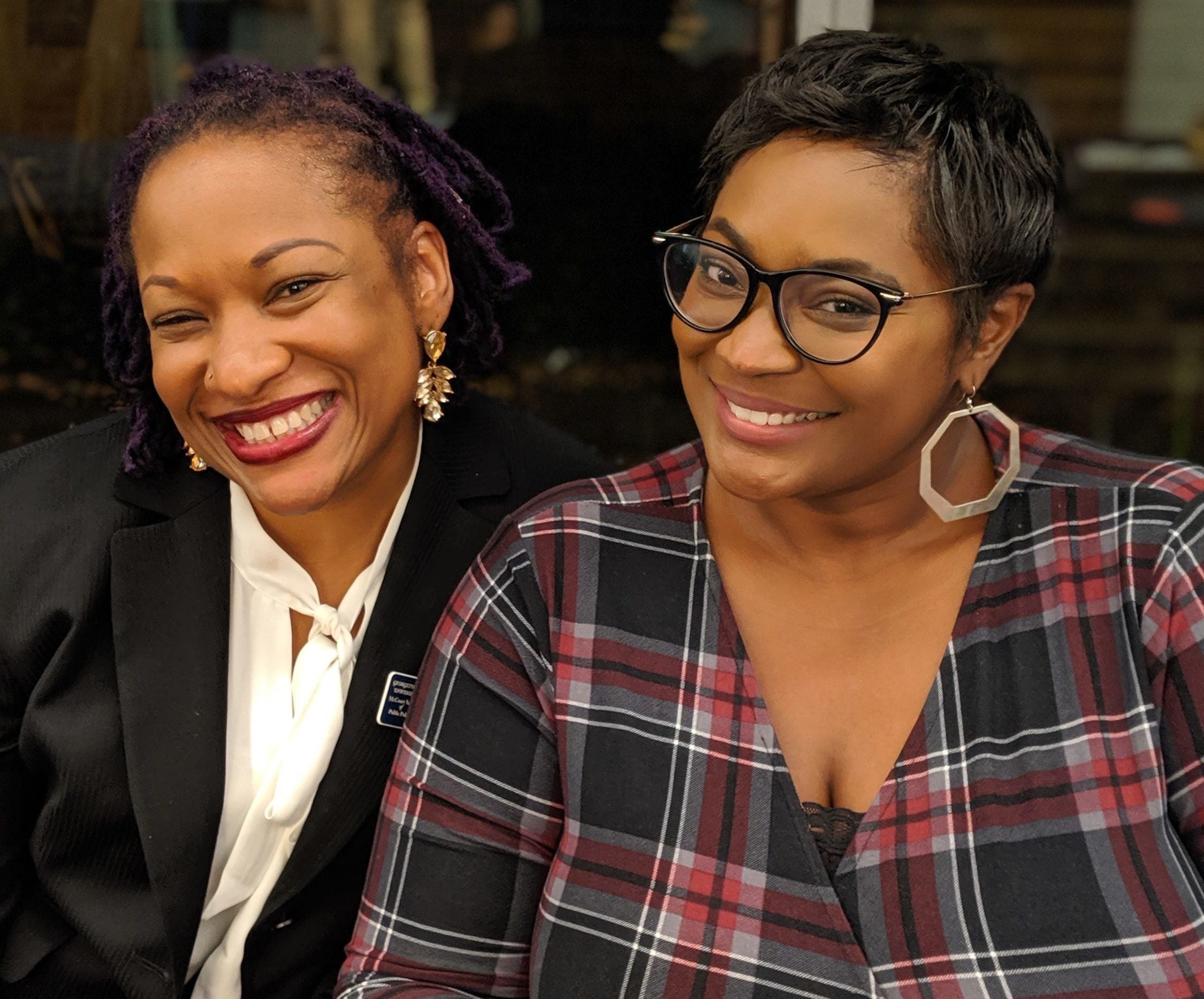 Two smiling women, one with purple hair and the other with short dark hair and glasses, pose closely together.