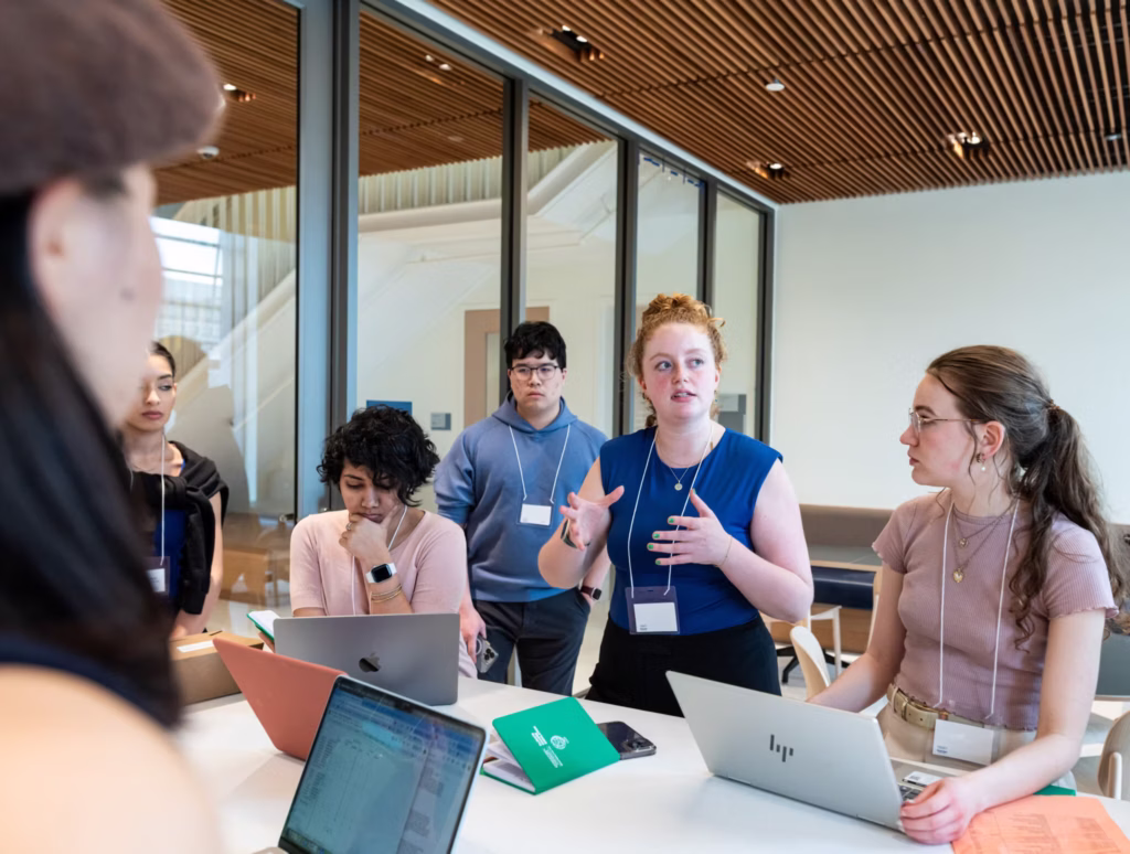 A group of six students working on a public policy project together, One student is talking with the group while the others work on their laptops or stand and listen.
