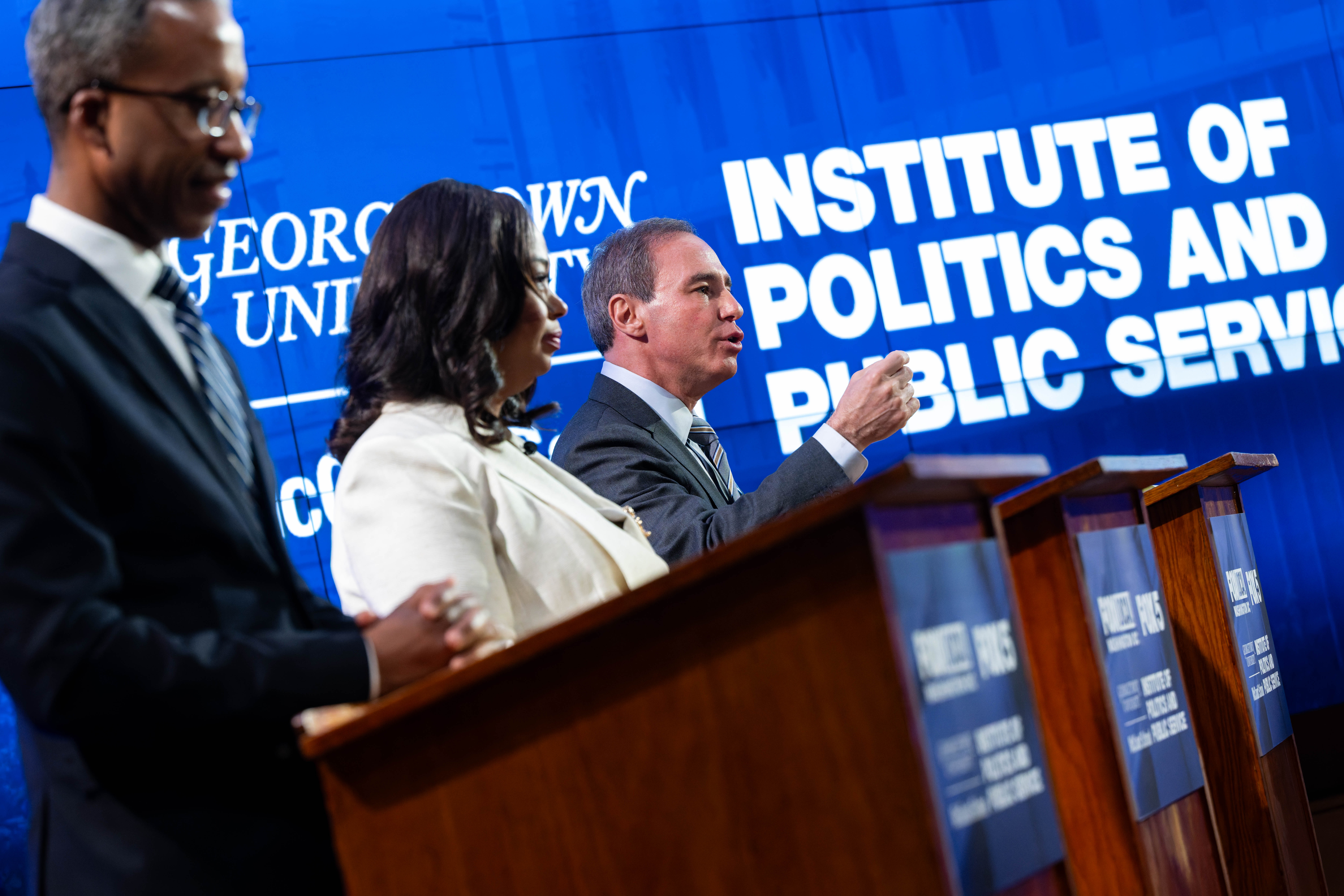 Three DC Mayoral candidates stand behind lecterns during debate, seen at profile with background text that reads "Institute of Politics and Public Service"