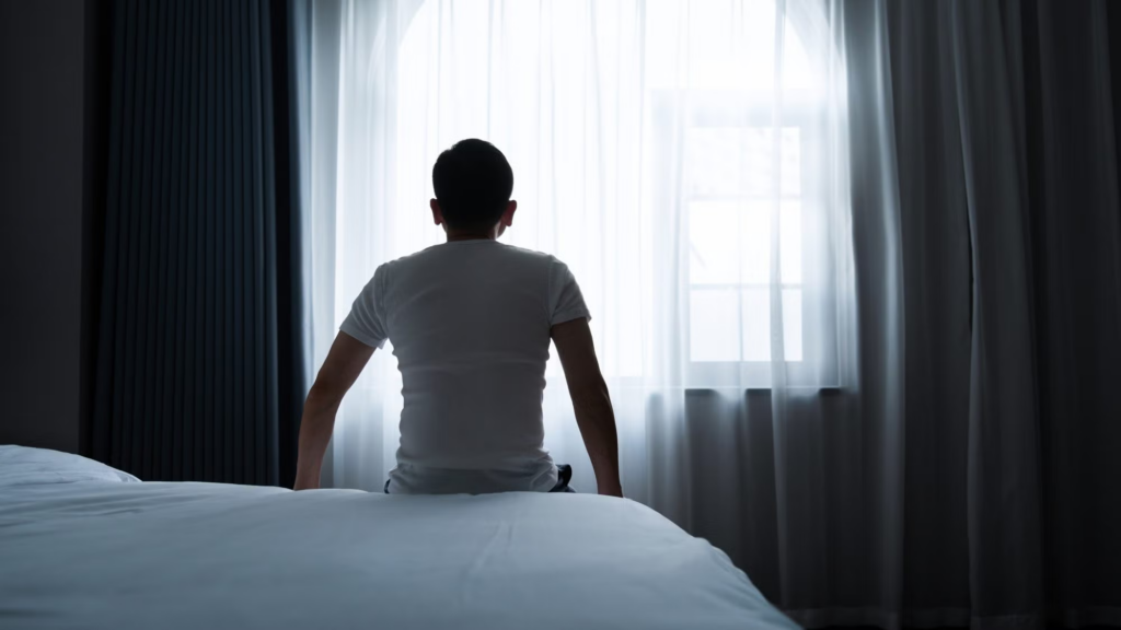 Individual sitting on bed looking through gloomy window.