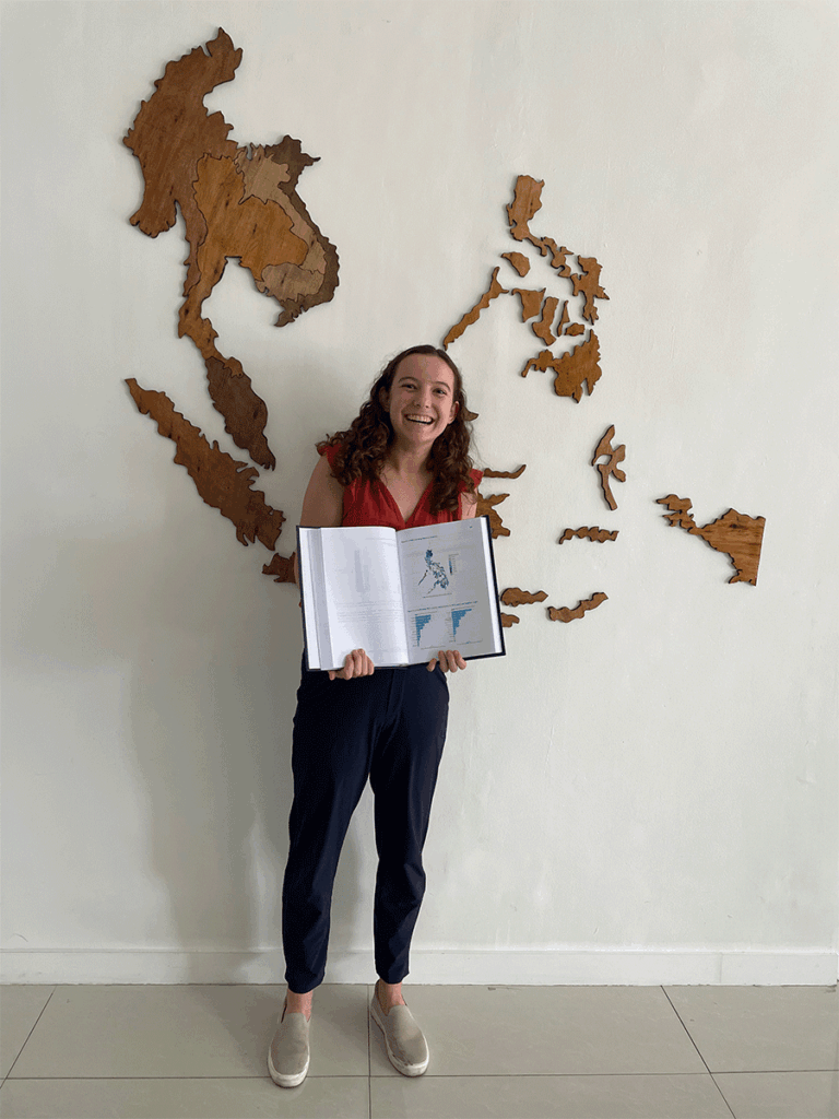 Hannah Reynolds posing in front of a map of the Southeast Asia region, holding booklet of personal research.
